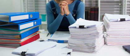 A woman clasps her hands at the center of two huge piles of paperwork.